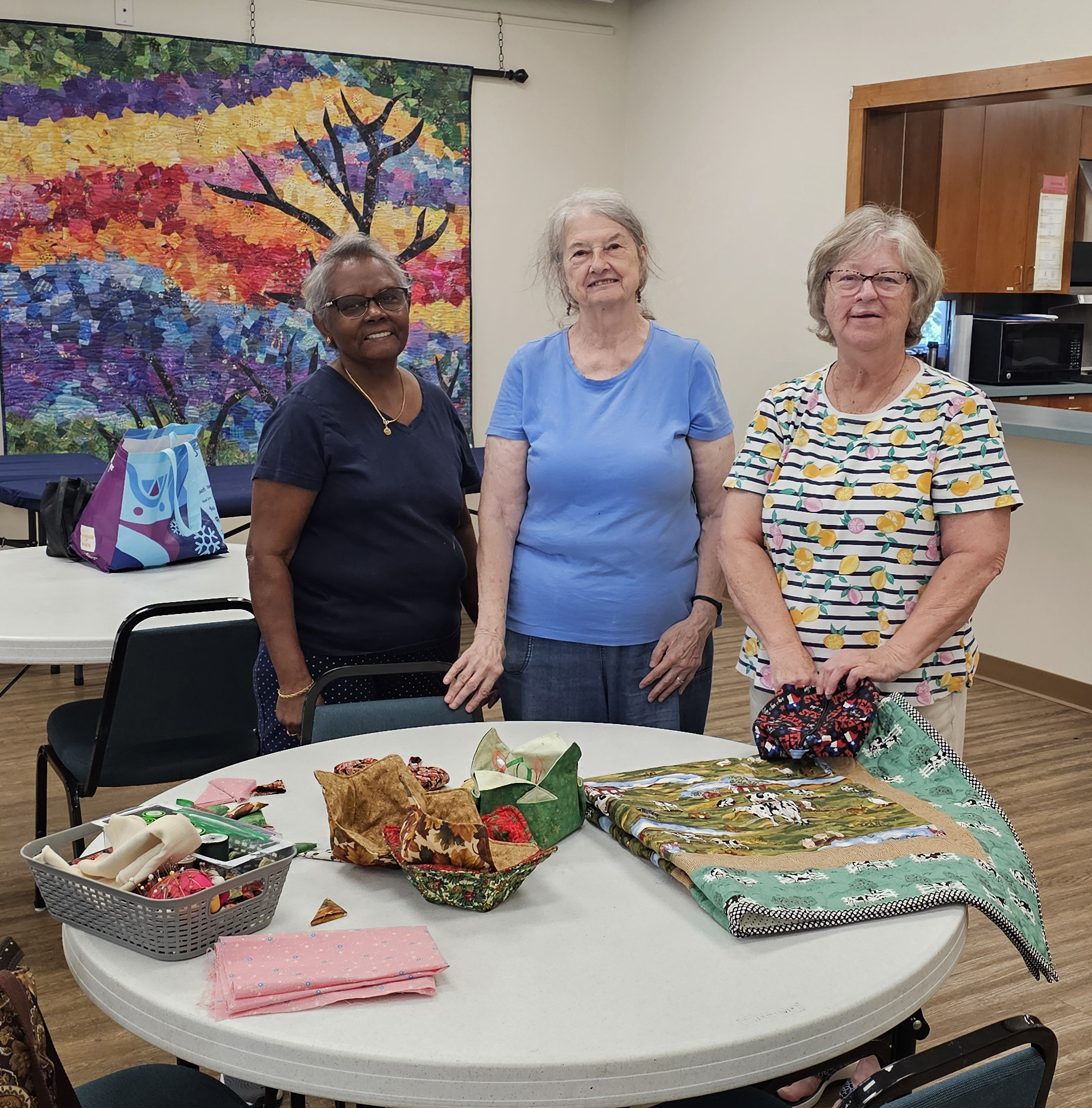 Pictured: Three members of the Trinity Stitchers ministry gathered around quilting supplies and an in-process quilt.